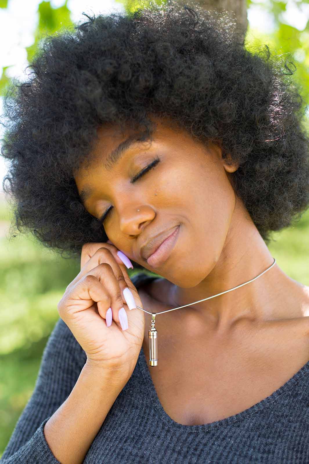 Young black woman wearing and holding up the beautiful Capsule Memorial Urn Necklace in Silver from online keepsake jewelry shop they made me wear it. Now you can safely insert the ashes of your beloved into the beautiful keepsake.