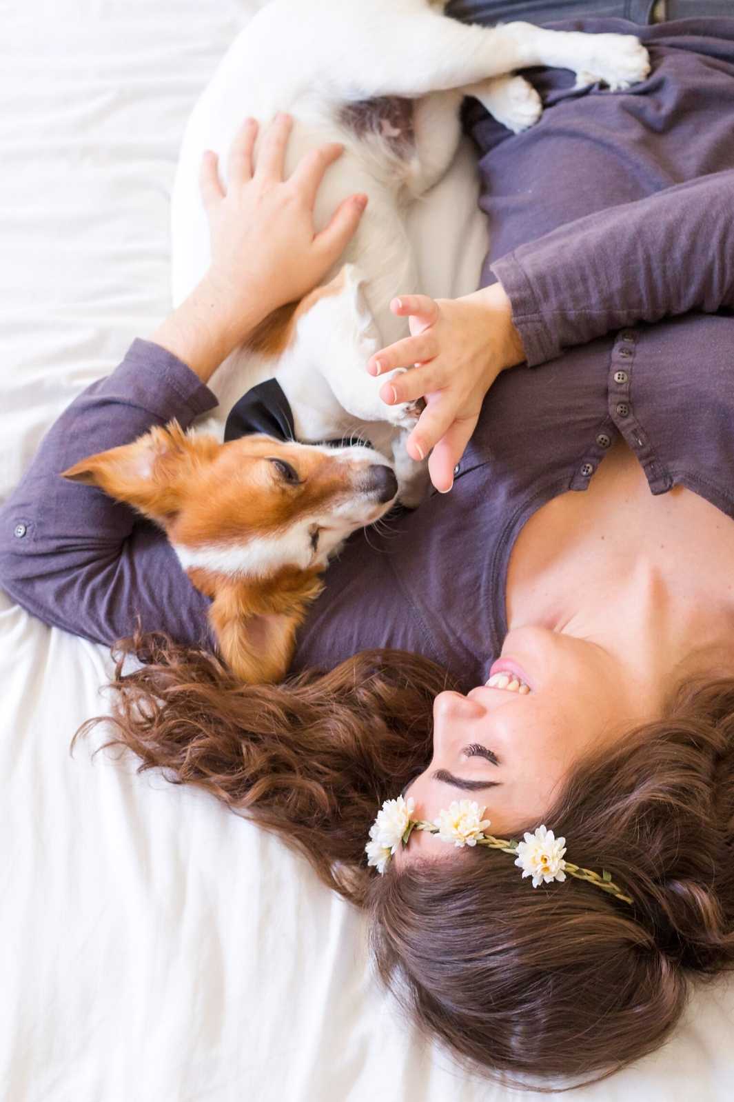 Young woman wearing a flower crown, lying on bed and cuddling with her cute small dog beside and celebrating National Hug Your Dog Day and National Pet Day!