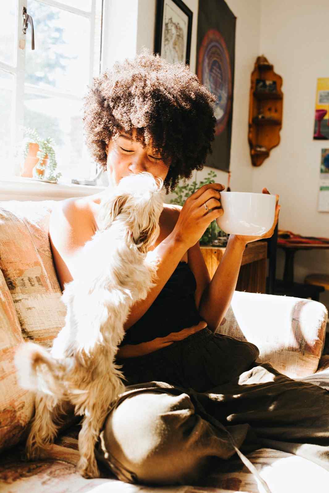 Young black woman drinking coffee on the couch and playing with her small dog, celebrating National Best Friends Day!