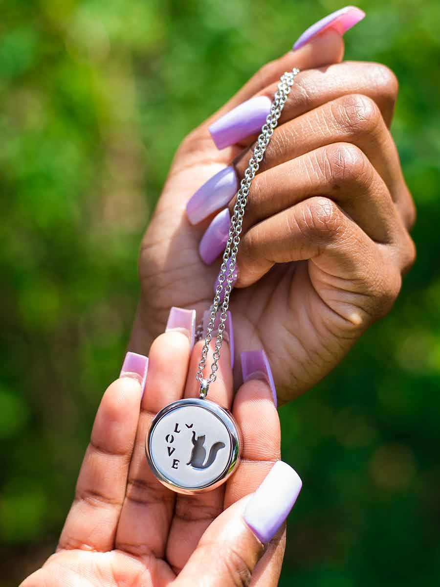 Black woman holding up silver Cat Memorial Locket. The perfect bereavement gift for family and friends who want a cremation locket to hold a small amount of pet ashes to remember their cat who crossed over the rainbow bridge. The locket can also be used as an aromatherapy essential oil diffuser as it comes with 12 free felt pads. Available from online boutique they made me wear it.