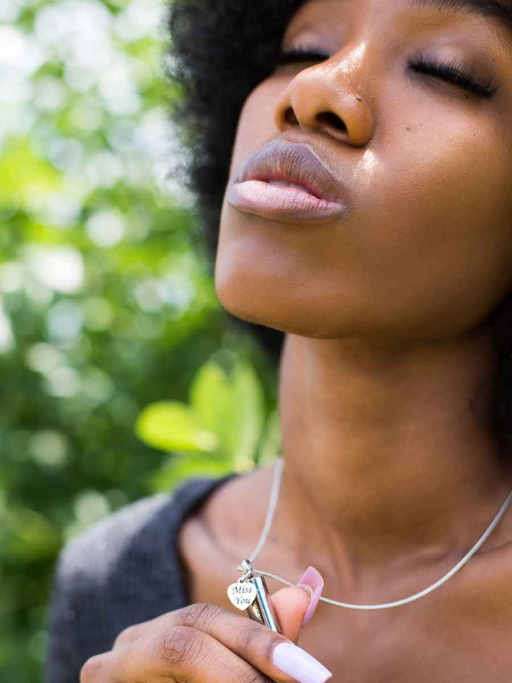 Black woman wearing and holding up the beautiful Miss You Memorial Urn Necklace with paw prints and a lovely heart-shaped tag engraved with the words "Miss You". The perfect bereavement gift for family and friends who want to wear an urn necklace to hold a small amount of pet ashes to remember their pet who crossed over the rainbow bridge. Available from online boutique they made me wear it.